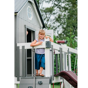 Child standing on a playhouse deck with a slide and telescope, surrounded by greenery.