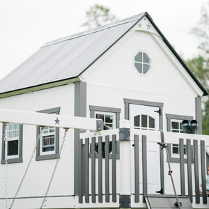 large White playhouse with gray accents and a swing set in front, surrounded by trees.