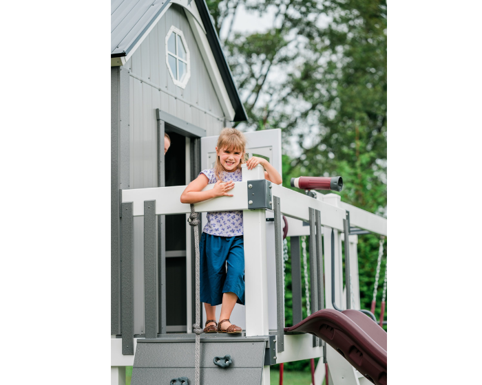 Child standing on a playhouse deck with a slide and telescope, surrounded by greenery.