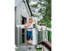 Child standing on a playhouse deck with a slide and telescope, surrounded by greenery.