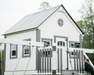 large White playhouse with gray accents and a swing set in front, surrounded by trees.
