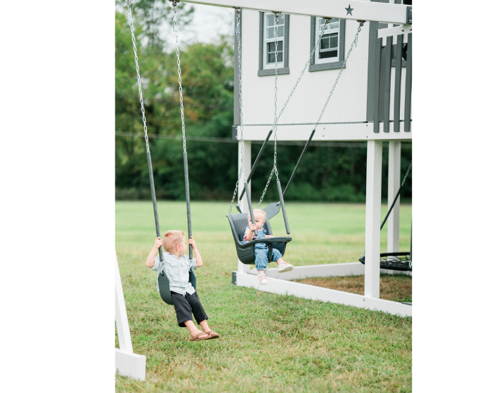 Two children on swings in front of a playhouse in a grassy area.